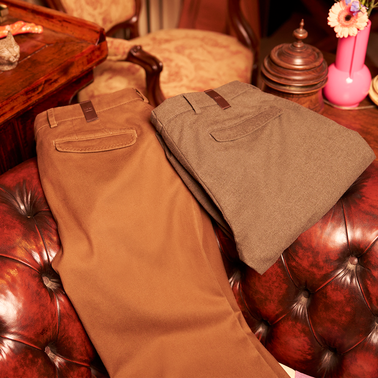 Two pairs of brown pants on a red leather couch with decorative items in the background.