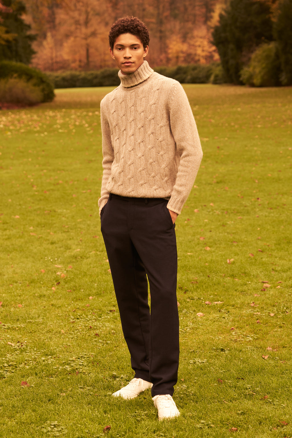 Man wearing a beige cable knit sweater and dark pants standing in a grassy field.