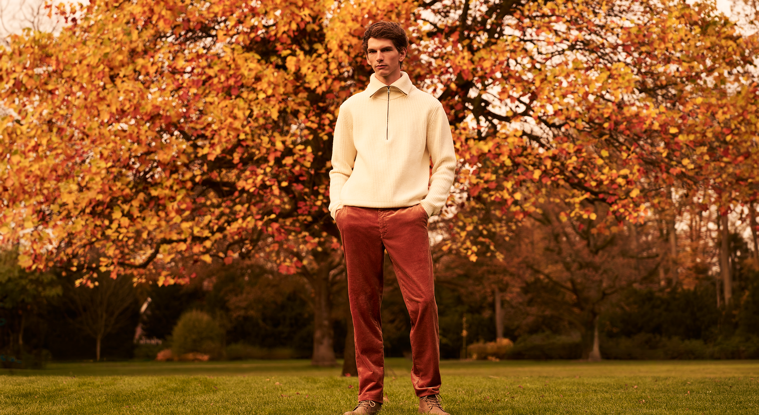 Man in a cream jacket and red pants standing in an autumn park.