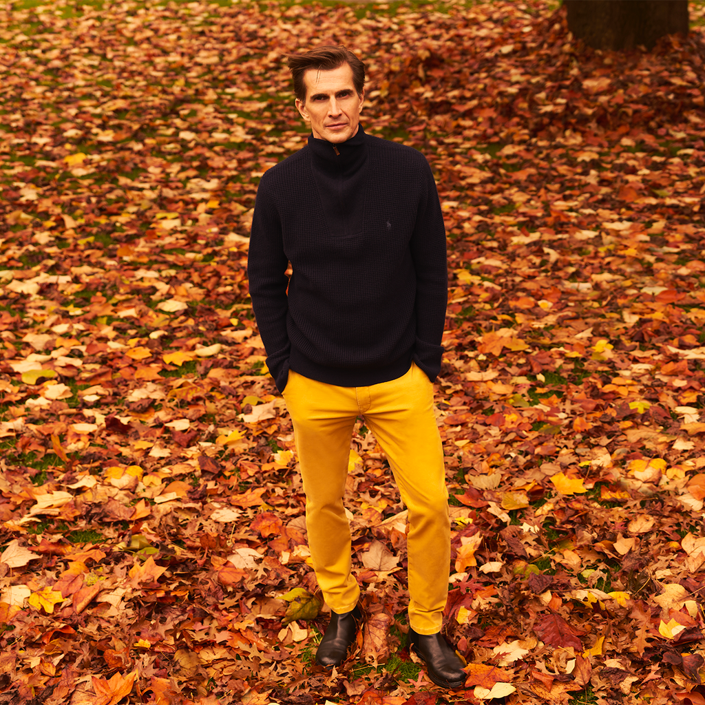 Man in a black turtleneck and yellow pants standing on a bed of autumn leaves.