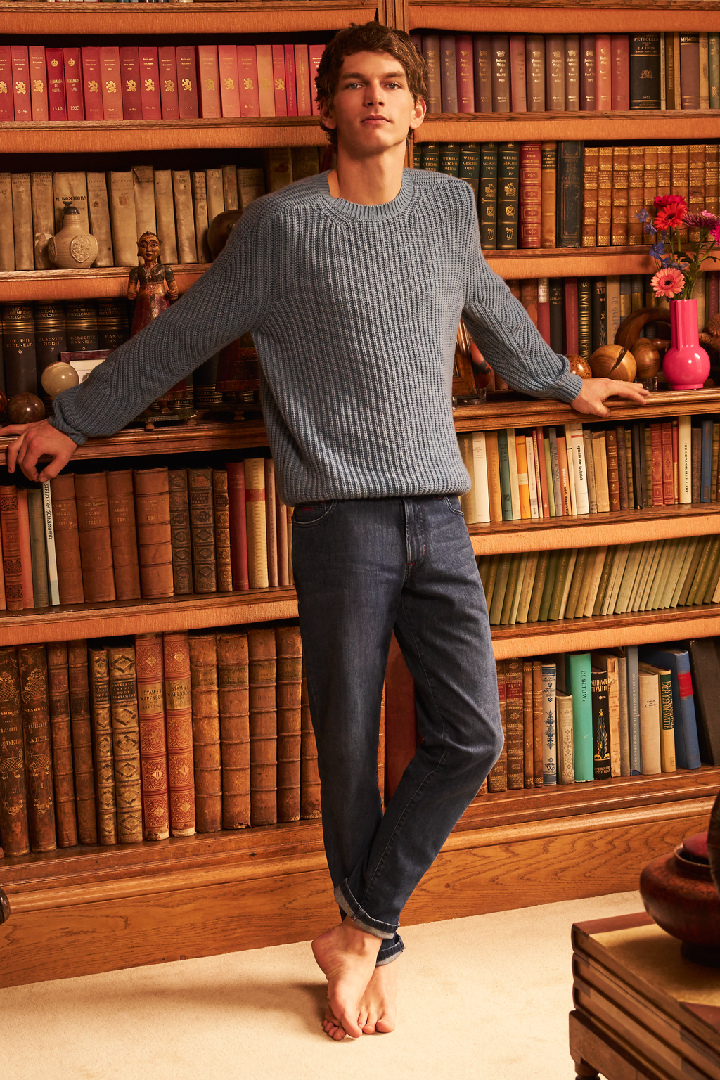 Man in a gray sweater and jeans standing in front of a bookshelf filled with books.